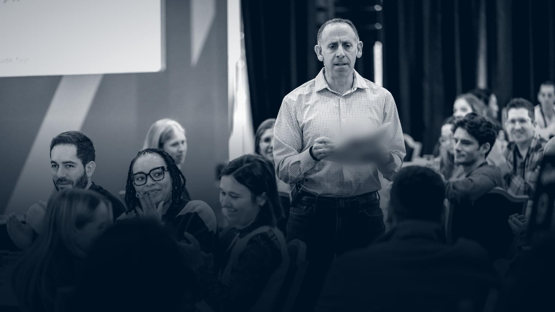 Russell standing among tables holding notes, the people around him laughing.