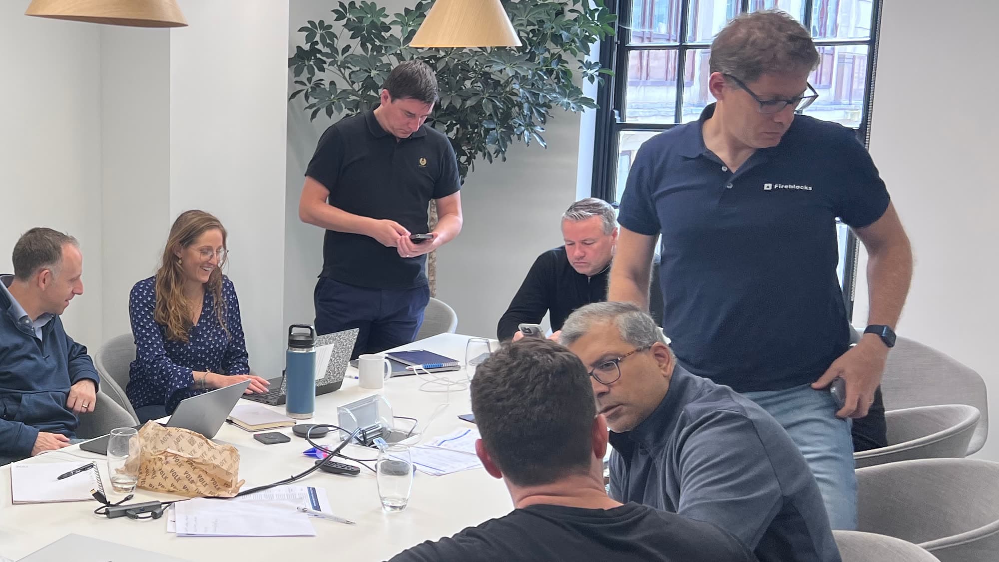 Seven people around a table in a small office, laptops and papers out, mid working session.